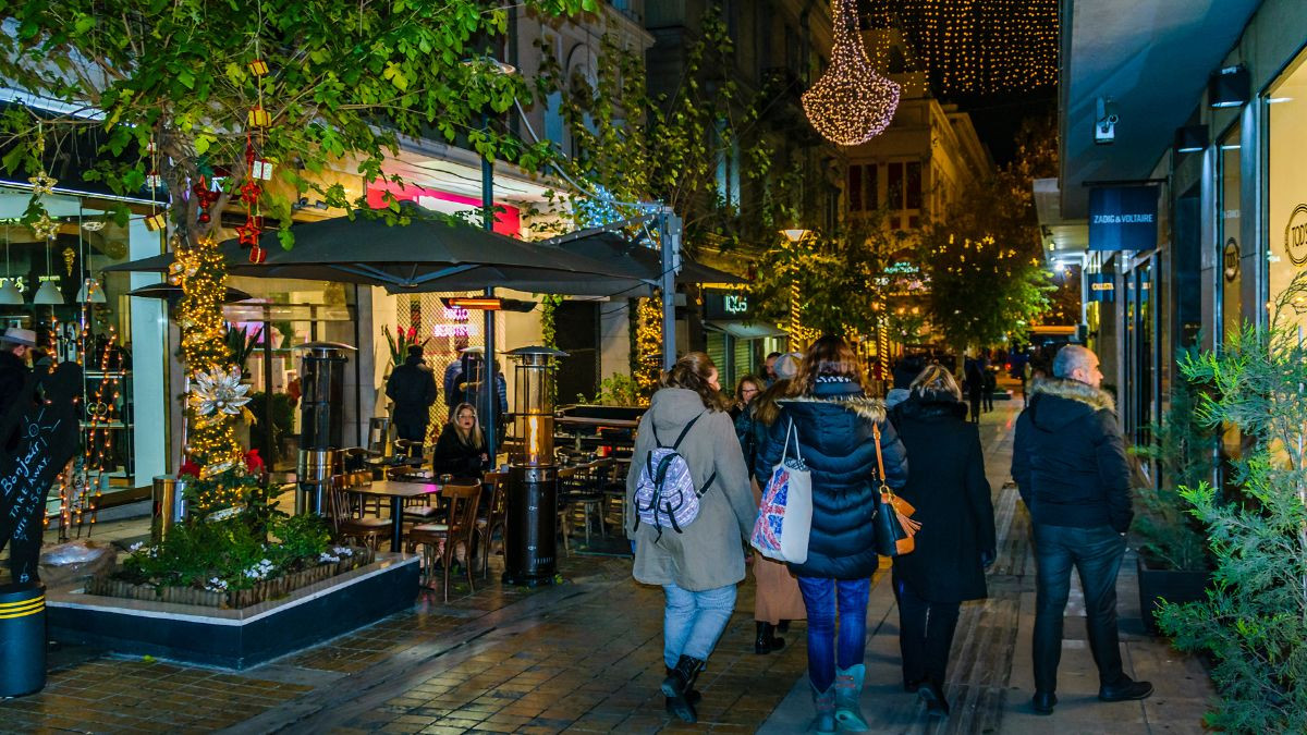 People walking on a festively decorated shopping street in Athens during Christmas time, with outdoor cafes and heaters, illuminated by warm lights and adorned with holiday decorations, creating a cozy and inviting atmosphere.
